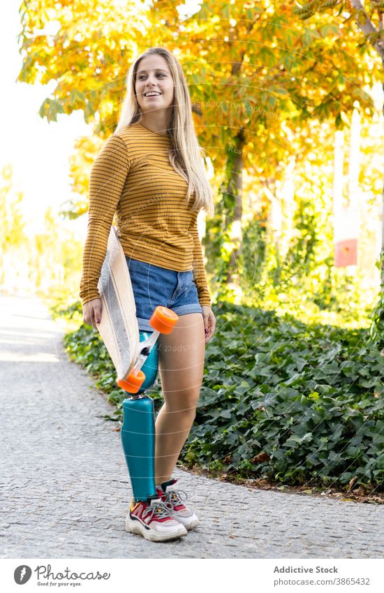 Smiling woman with longboard walking on street leg prosthesis bionic skater artificial limb disable urban female skateboard amputee town modern summer slim