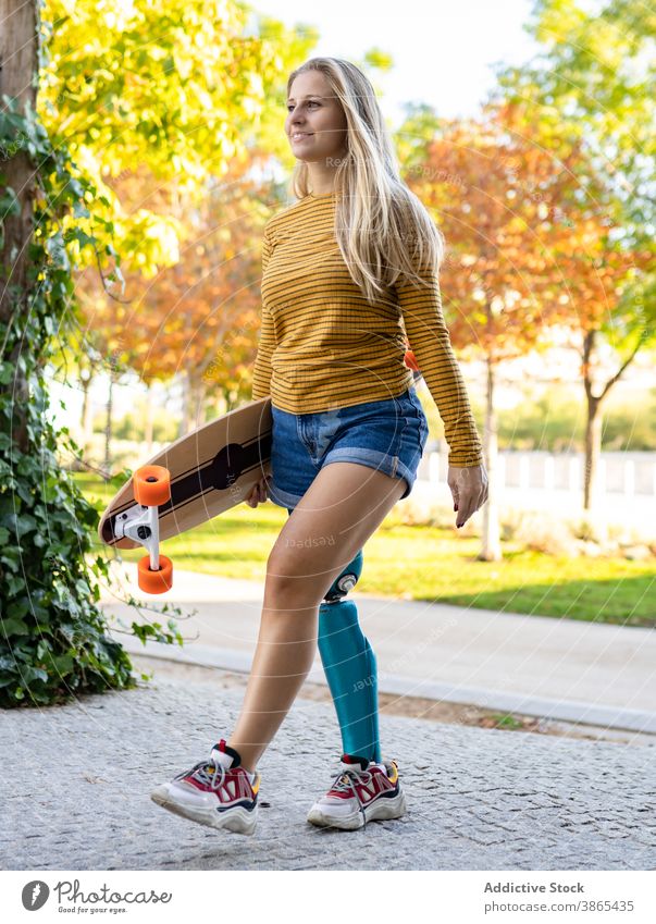 Smiling woman with longboard walking on street leg prosthesis bionic skater artificial limb disable urban female skateboard amputee town modern summer slim