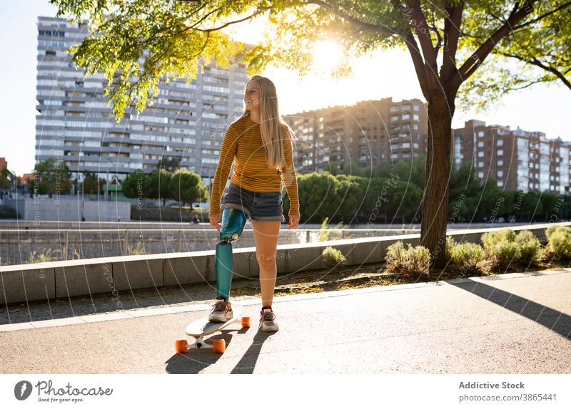 Content woman with longboard in park leg prosthesis bionic skater artificial limb urban summer female cheerful amputee disable street happy optimist town