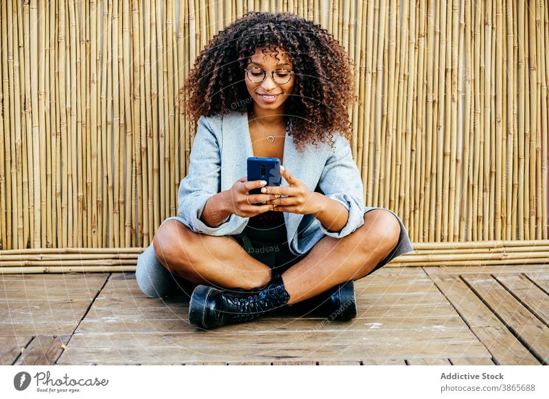 Cheerful black woman using smartphone near bamboo wall rest style floor wooden smile legs crossed female young outfit weekend trendy ethnic african american