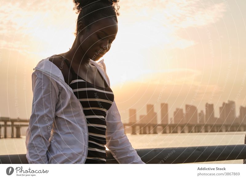 Calm black woman standing on city embankment at sunset sea tranquil pensive relax calm sunlight urban young female african american ethnic seafront thoughtful