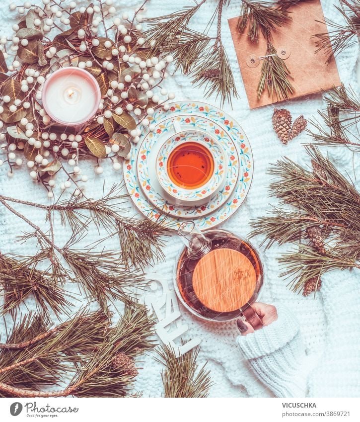 Winter and Christmas lifestyle. Woman hand holding tea pot on white knitted blanket with cup of tea, Christmas wreath and fir cones and branches. Top view