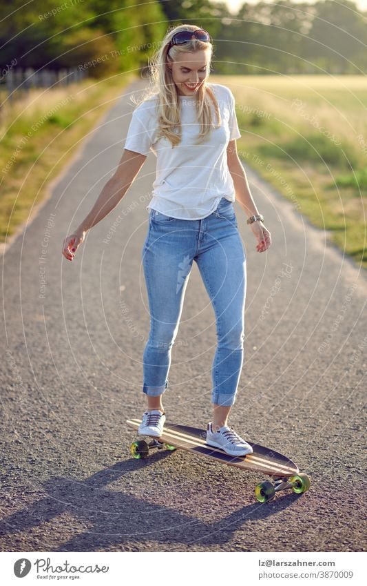 Fit active middle-aged woman playing on her skateboard approaching the camera along a narrow rural road with a happy smile backlit by the evening sun fit blond