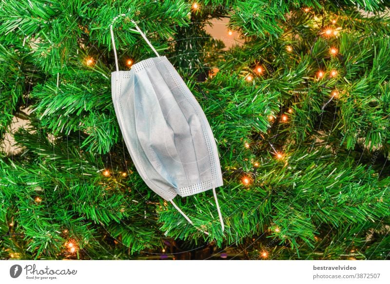 Christmas covid-19 mask hanging as a decoration on a green tree with lights. Coronavirus face protection plain design used as seasonal ornament at the branches of an artificial tree.