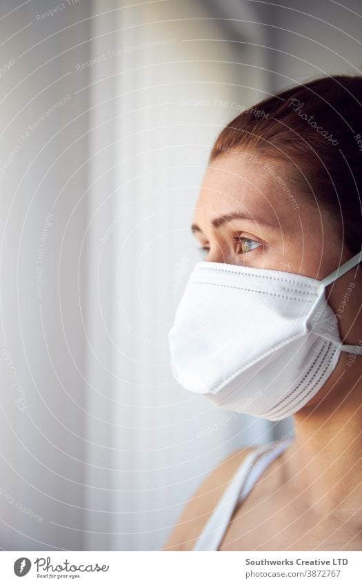 Young woman looking out of window wearing protective face mask. female home virus disease diseases flu air health sick prevention inside outside isolation self