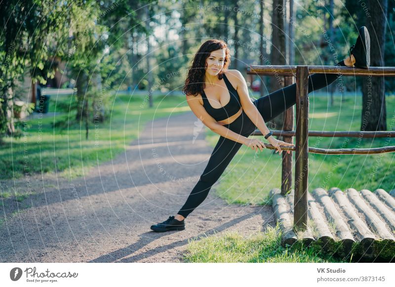 Flexible young curly woman wears black top leggings and sneakers, stretches her legs, does sports in the fresh air on summer days, poses in front of green trees. People, flexibility and exercise concept