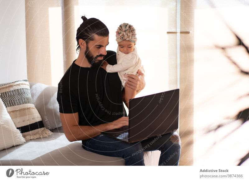 Father using laptop while sitting on sofa with baby multitask father work home freelance busy toddler cute ethnic couch adorable browsing online apartment