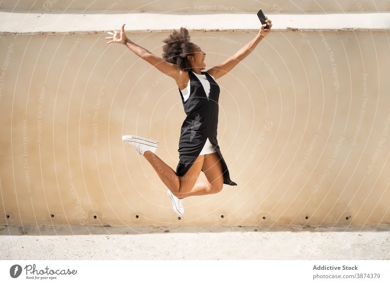 Positive ethnic woman jumping on street with smartphone carefree selfie having fun freedom outstretch excited moment female black african american gadget device