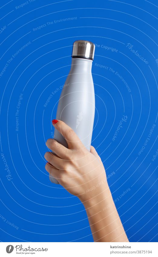 Close up of female hand, holding grey reusable steel bottle on blue background monochrome ecologic water thermo bright mockup woman faceless aluminum blank