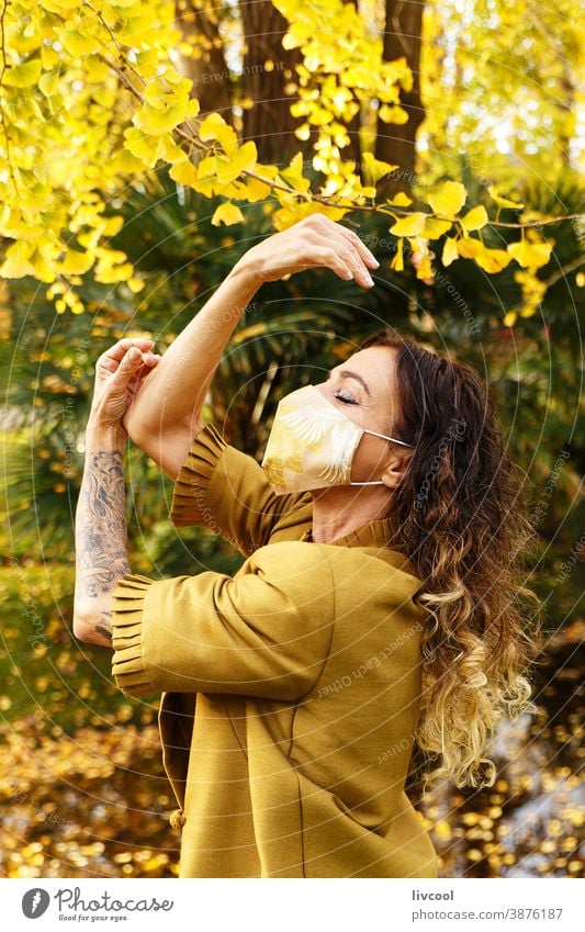 woman in yellow dress and mask in a park retouching hair garden yellowish leafs lifestyle mature portrait one people tree coat yellow overcoat scene romantic
