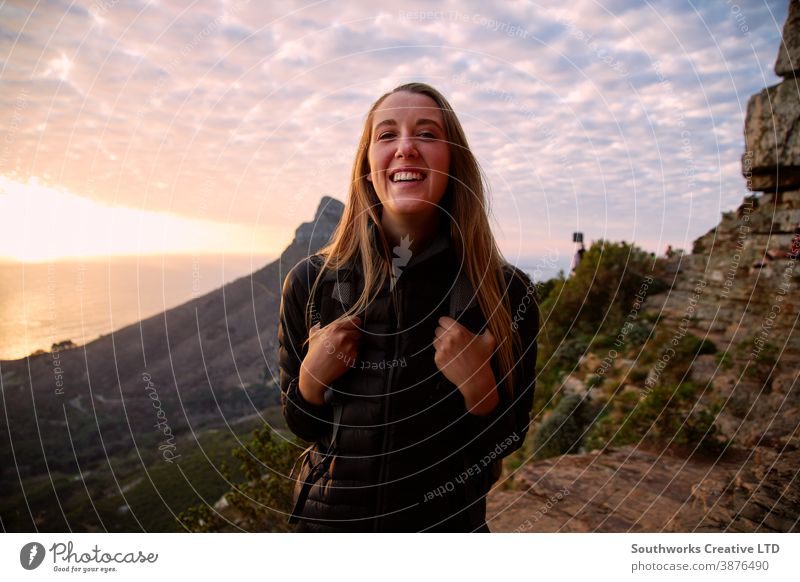 Portrait Of Smiling Young Woman Hiking Along Coastal Path As Sun Sets Over Sea Behind Her travel vacation happy woman young women hike hiking walk walking trek