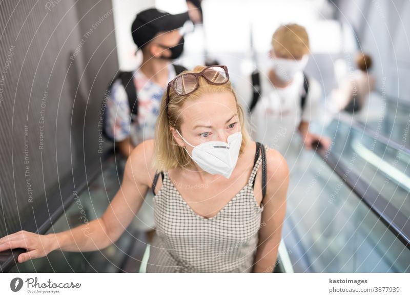 Portrait of casual yound woman uses escalators in the department store wearing protective mask as protection against covid-19 virus. Incidental people on the background
