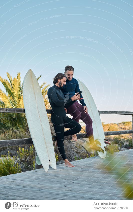 Cheerful surfers using smartphone together on embankment promenade surfboard seaside cheerful gadget boardwalk device summer friend friendship browsing