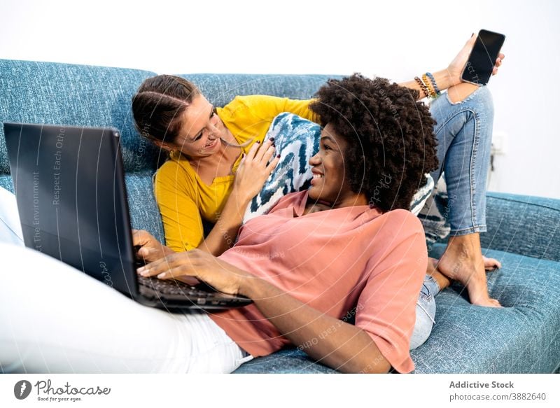 Cheerful multiracial lesbian couple with gadgets chilling on sofa women at home rest having fun together relationship love girlfriend happy affection young