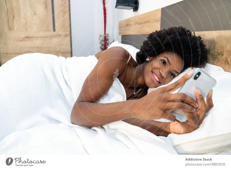 Black woman using smartphone in bed browsing morning awake smile addict social media female ethnic black african american mobile home internet bedroom message