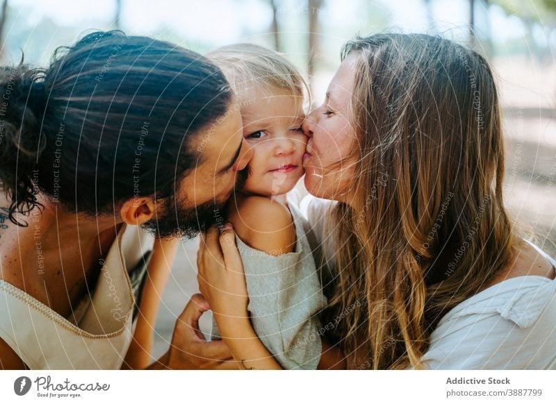 Mother and father kissing kid together family cheek child love mother cuddle unity multiethnic multiracial diverse forest hug tender daughter parent happy