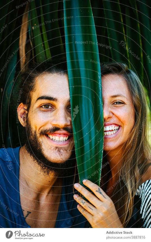 Positive couple in tropical garden looking at camera having fun cheerful hide plant leaf laugh entertain playful multiethnic multiracial diverse exotic park