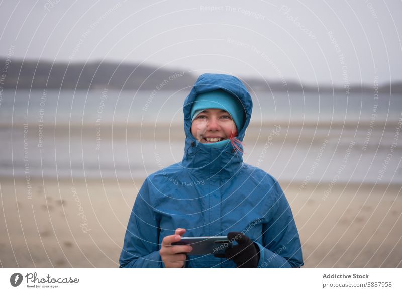 Cheerful traveling woman with smartphone near sea in autumn traveler explorer warm clothes wind seashore cheerful female waterproof jacket cellphone seaside
