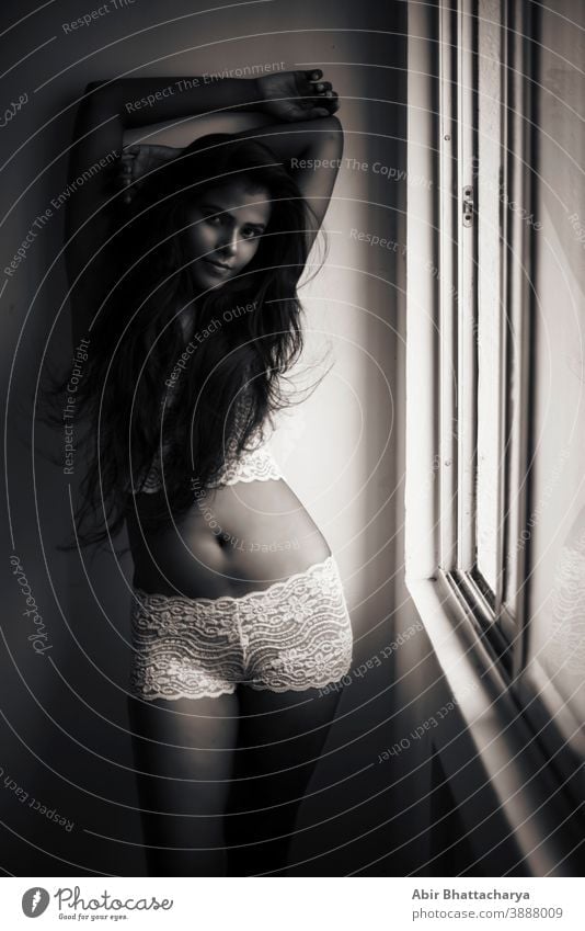 Portrait of a young and attractive dark skinned Indian Bengali woman in lingerie posing in a casual mood in front of a window. Boudoir photography. adult art