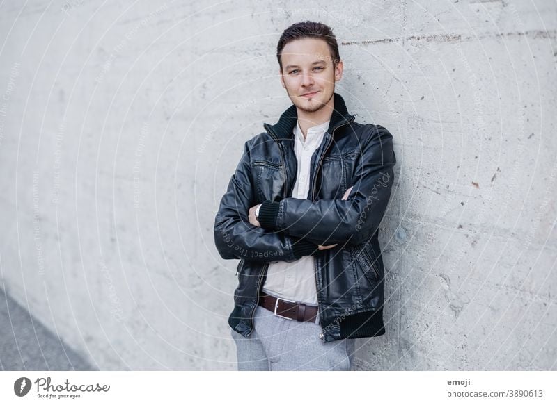 young man in leather jacket, business, outside in front of concrete wall masculine more adult Upper body arms folded Young man Man Friendliness Smiling Positive