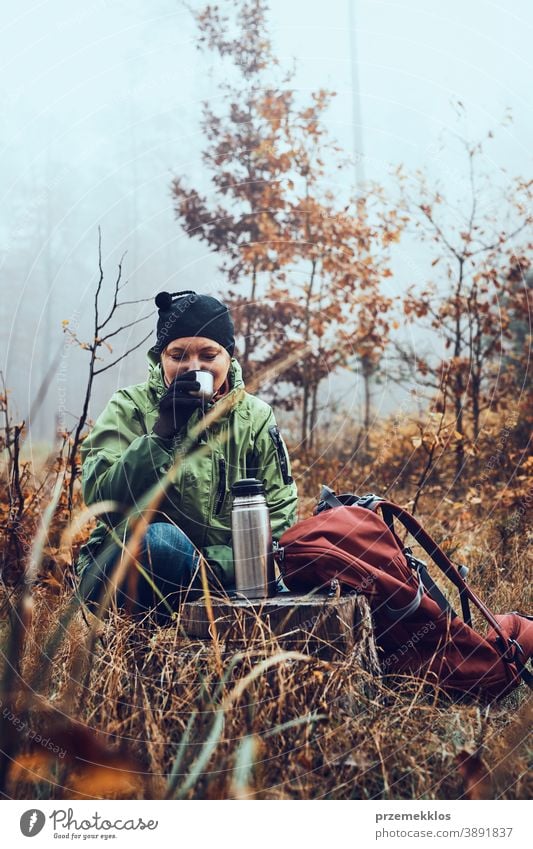 Woman with backpack having break during autumn trip drinking a hot drink from thermos flask on autumn cold day outdoors destination hiking holiday vacation