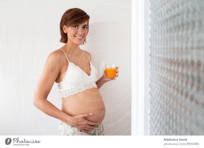 Smiling pregnant woman with glass of juice at home healthy drink pregnancy fresh orange female bra expect belly maternal charming anticipate lingerie smile