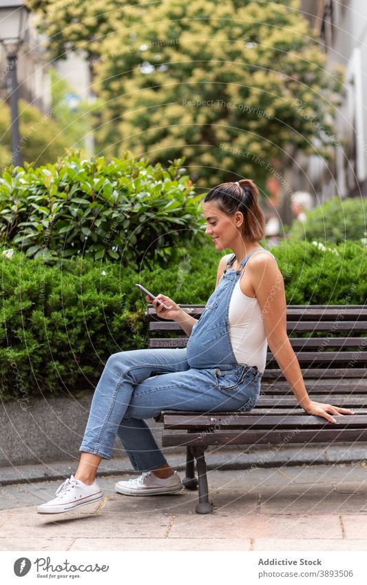 Smiling pregnant woman using smartphone in city browsing street female bench wooden gadget calm surfing connection online communicate abdomen parent await