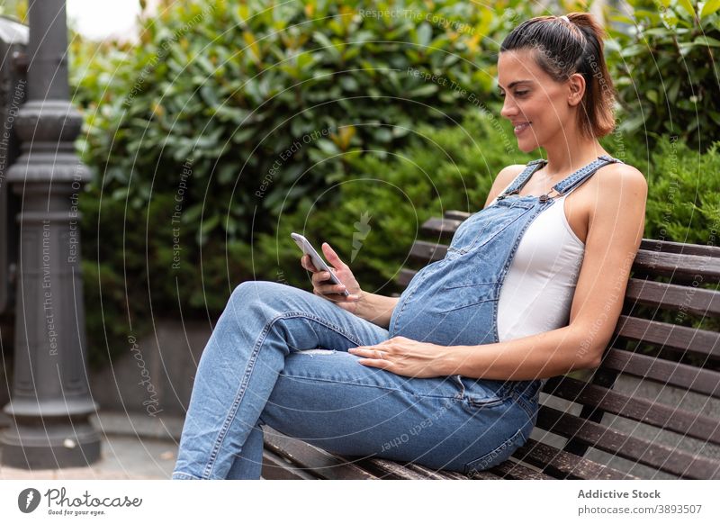 Smiling pregnant woman using smartphone in city browsing street female bench wooden gadget calm surfing connection online communicate abdomen parent await
