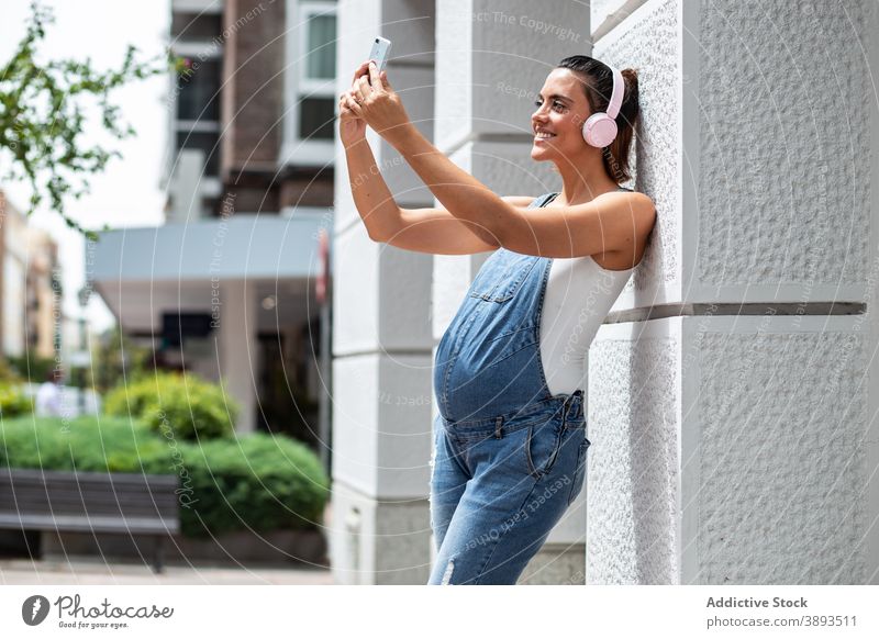 Delighted pregnant woman taking selfie in city pregnancy stroll headphones music self portrait cheerful female street urban building photo mobile happy smile