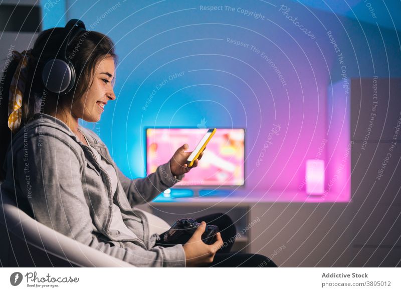Smiling female gamer using smartphone at home woman gamepad controller console browsing joystick videogame cheerful evening smile headphones entertain sit