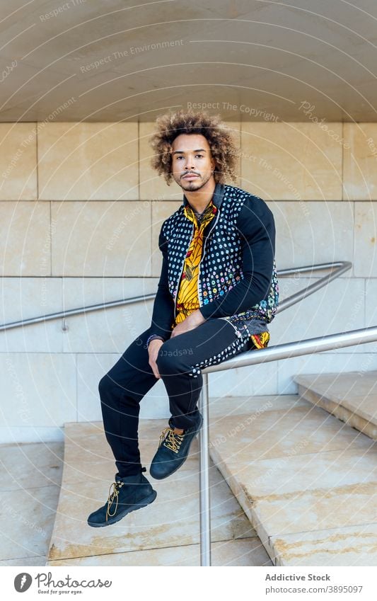 Stylish hipster ethnic man sitting on railing style trendy informal fashion curly hair beard young modern outfit step guy african american black male lifestyle