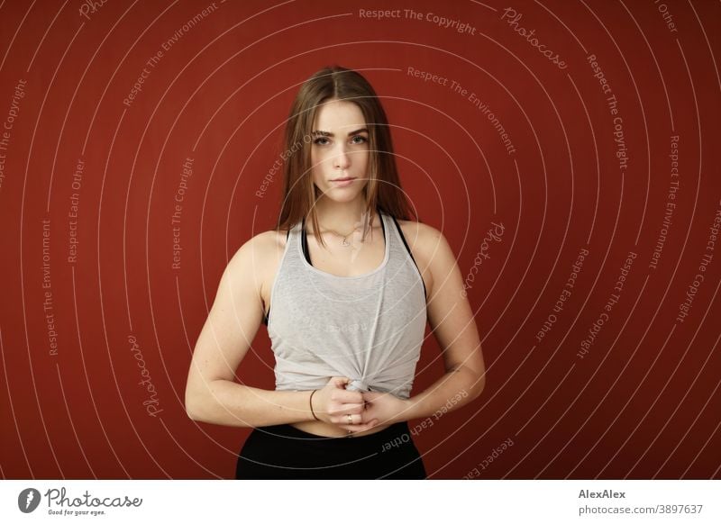 Portrait of a young, sporty woman with a six-pack in sports clothes in a room in front of a red wall Student daintily Jewellery Facial expression empathy