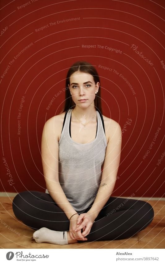 Portrait of a young, sporty woman in sports clothes in a room in front of a red wall Student daintily Jewellery Facial expression empathy