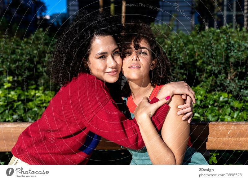 Smiling lesbian couple embracing and relaxing on a park bench affectionate closeness ethnic ethnicity friends friendship laughing lover loving multiracial