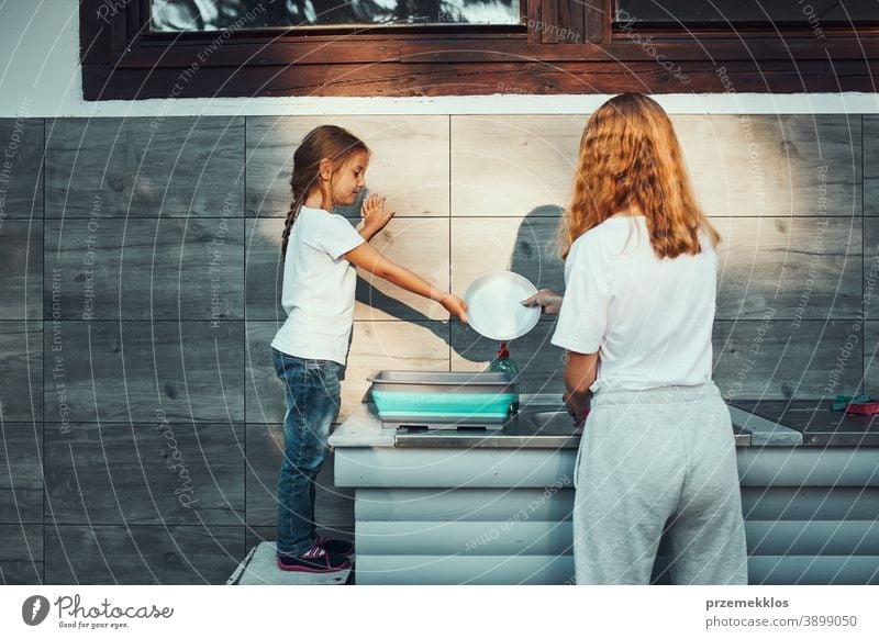 Teenager girl washing up the dishes pots and plates with help her younger sister in the outdoor kitchen during vacations on camping working together siblings
