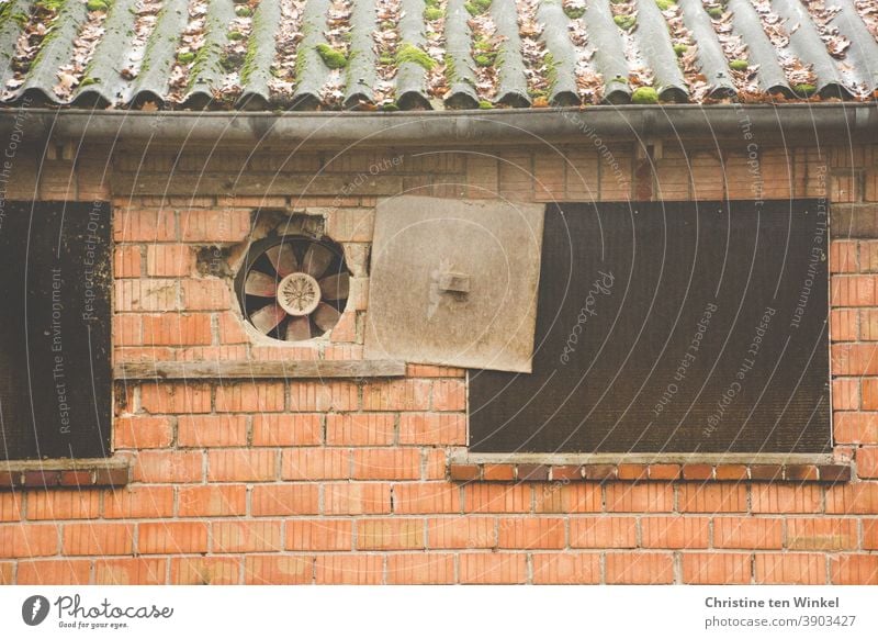 contemporary history | Fan in the outer wall of an old agricultural building with a tin roof. The cover plate has been pushed aside. Some stones are beginning to crumble away. The window openings next to it are closed with wooden boards.