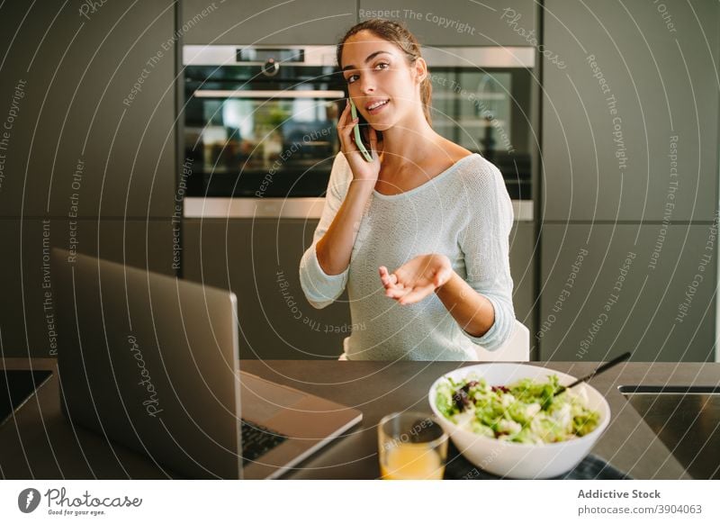 Woman talking on smartphone in kitchen woman at home laptop remote check mobile message young female gadget device internet browsing online communicate