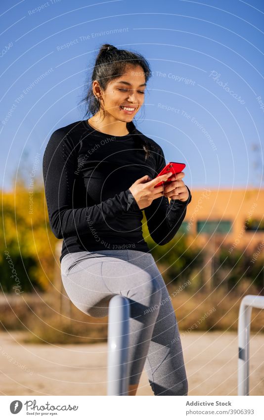 Smiling ethnic sportswoman browsing smartphone on sports ground break training calisthenics parallel bar relax surfing female sportswear athlete fitness device
