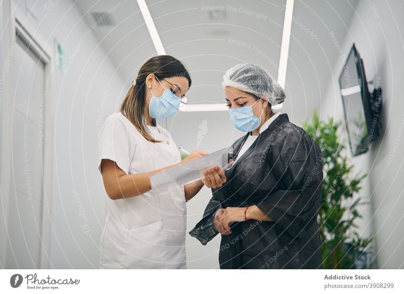 Female beauticians in uniform standing in corridor of clinic cosmetology women cosmetician together beauty dermatology clipboard take note specialist mask