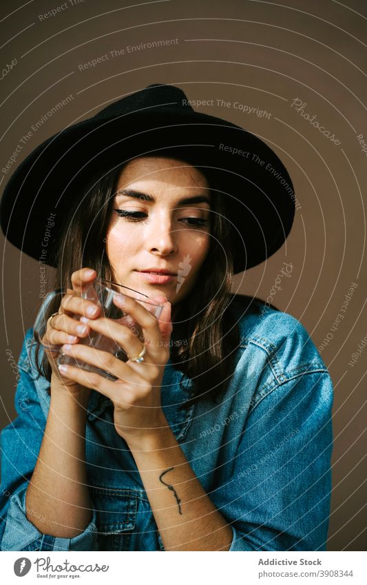 Stylish woman with empty glass in studio denim outfit hat trendy apparel style female sit young charming dreamy fashion relax ponder contemplate appearance