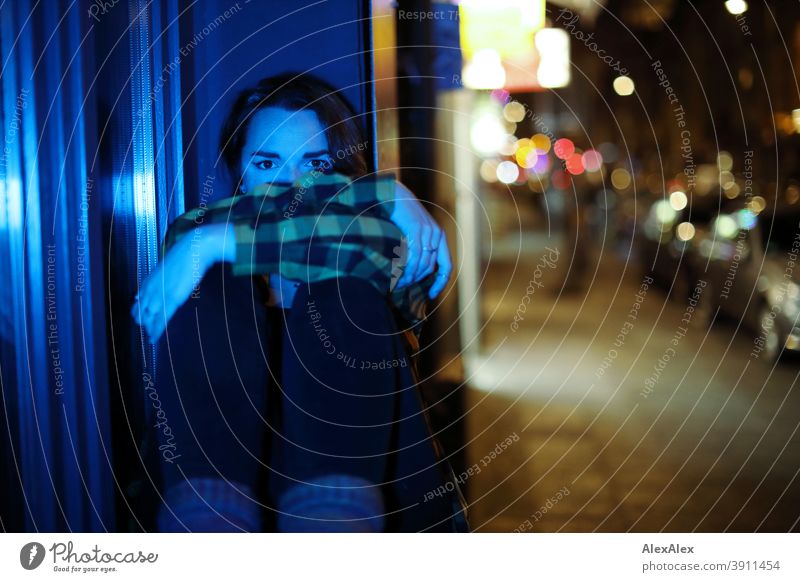 Junge Frau sitzt nachts blau beleuchtet in einem Fenster auf einer bunt beleuchteten Strasse jung Jugendliche blaues Licht Straße Karohemd kariert draussen