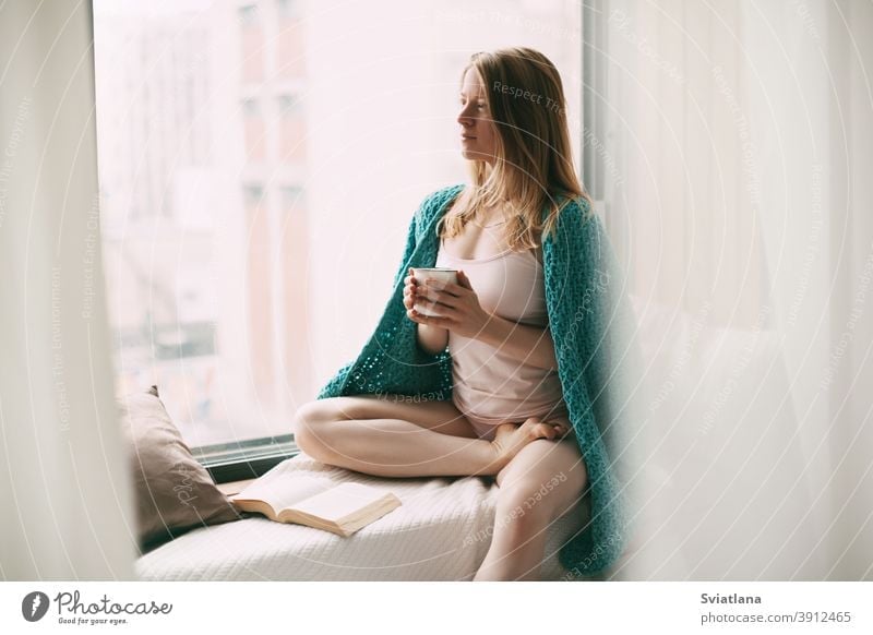 A young girl is sitting on the windowsill in the early morning, drinking hot coffee, and an open book is lying next to her woman leisure read home cup people
