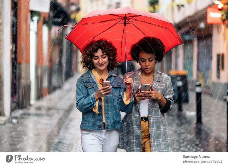 Happy Friends Using Phone umbrella rainy day friends afro girl black woman caucasian using phone city life smiling front view portrait women looking