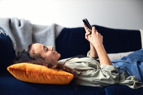 Young Smiling Woman Relaxing At Home Lying On Sofa Checking Social Media On Mobile Phone woman at home lying sofa lounge mobile mobile phone cell cell phone