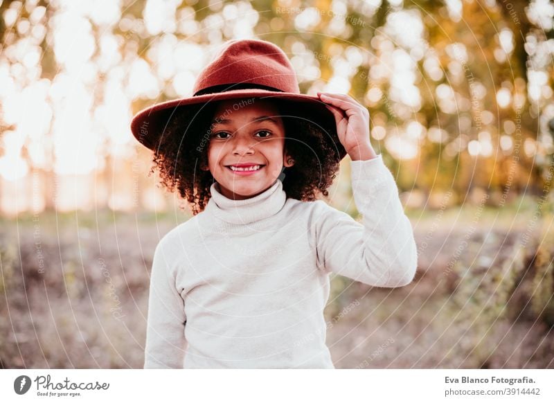 portrait of cute afro kid girl wearing a hat at sunset during golden hour, autumn season, beautiful trees background nature outdoors brown leaves