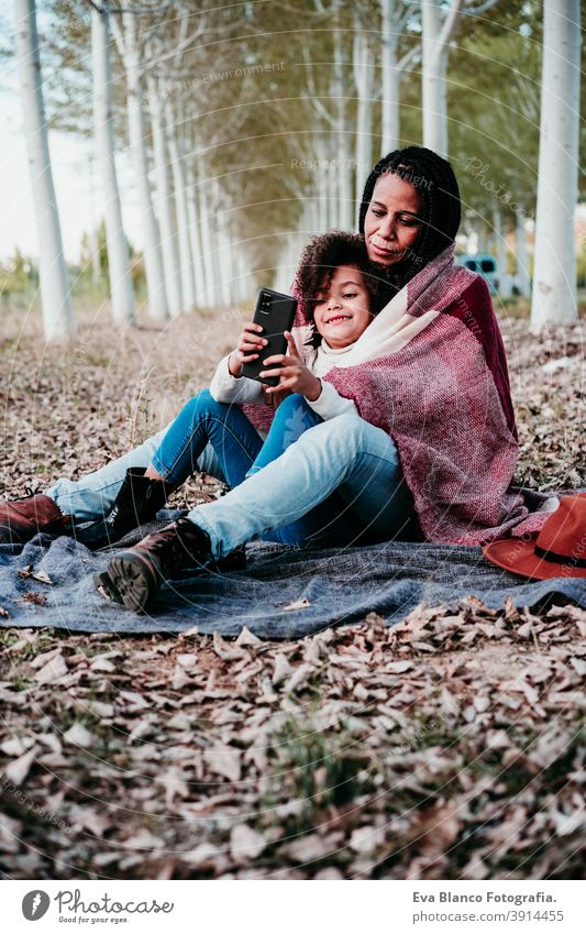 hispanic mother and afro kid girl outdoors hugging at sunset wrapped in blanket. Autumn season. Family and love concept MOBILE PHONE technology internet device