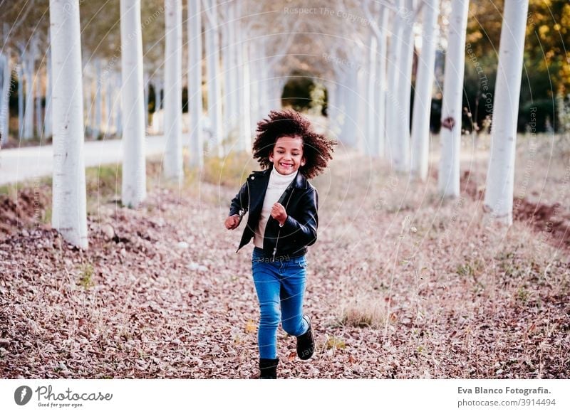 portrait of cute afro kid girl running at sunset during golden hour, autumn season, beautiful trees background fun nature outdoors hat brown leaves