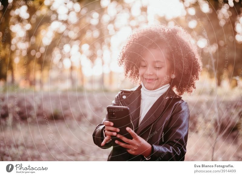 portrait of cute afro kid girl using mobile phone at sunset during golden hour, autumn season, beautiful trees background technology nature outdoors hat brown