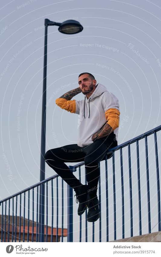 Hipster man sitting on fence on street hipster urban tattoo casual hoodie beard modern style male adult lifestyle rest cool trendy young subculture brutal