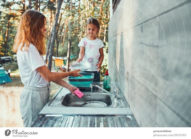 Teenager girl washing up the dishes pots and plates with help her younger sister in the outdoor kitchen during vacations on camping working together siblings